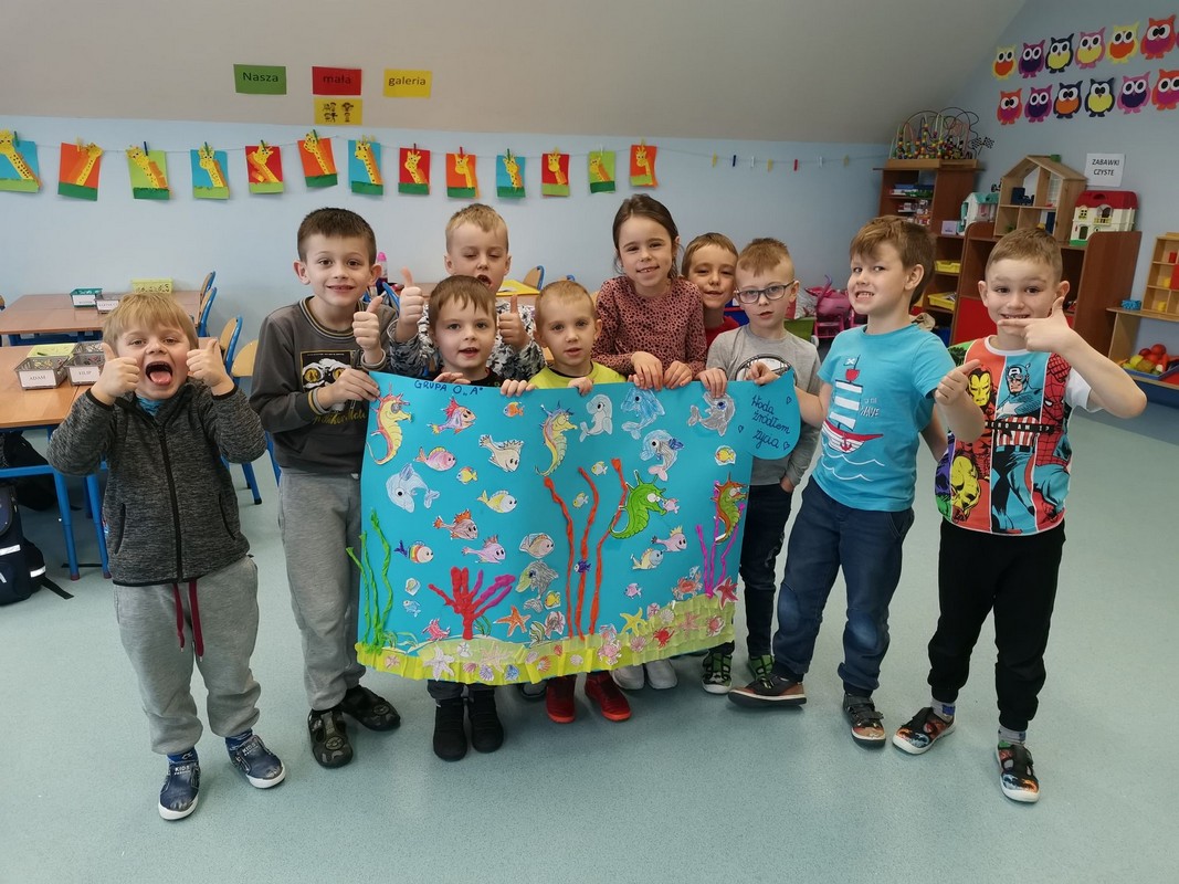 grupa dzieci stoi w sali przedszkolnej i trzyma duży plakat, na którym znajduje się wyklejona makieta dna oceanu. Jest bardzo kolorowa. Są tam i roślinki i kolorowe rybki.