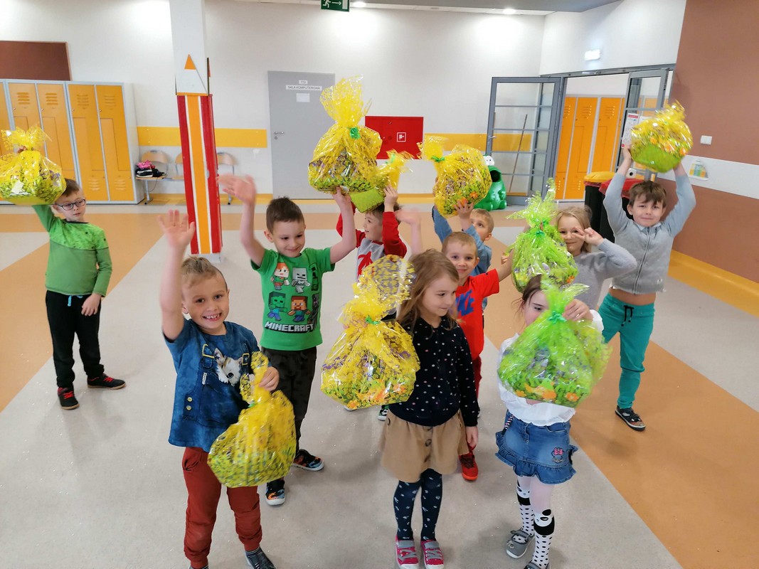 grupa dzieci kolorowo ubranych w żółtym holu szkoły z zielonymi paczkami od zajączka. Dzieci cieszą się.