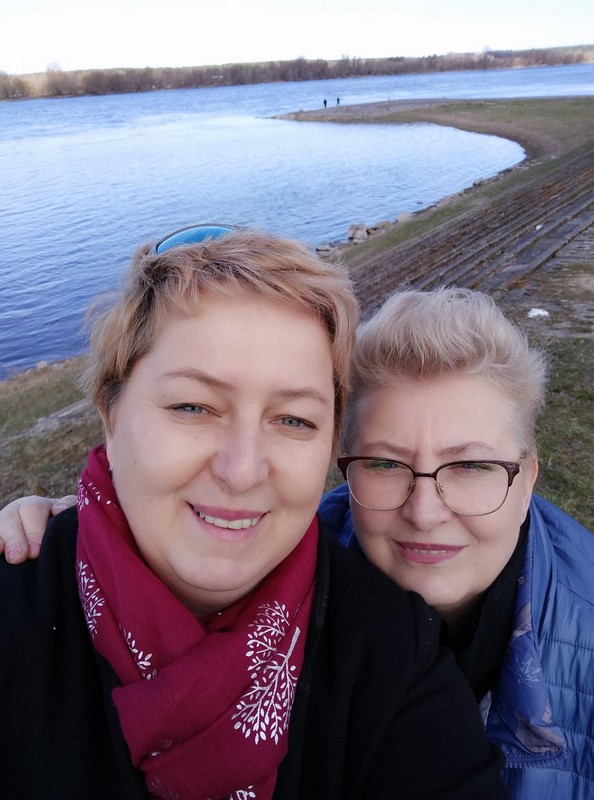 selfie dwóch krótkowłosych, uśmiechniętych blondynek nad brzegiem jeziora. Jedna z pań ma okulary  