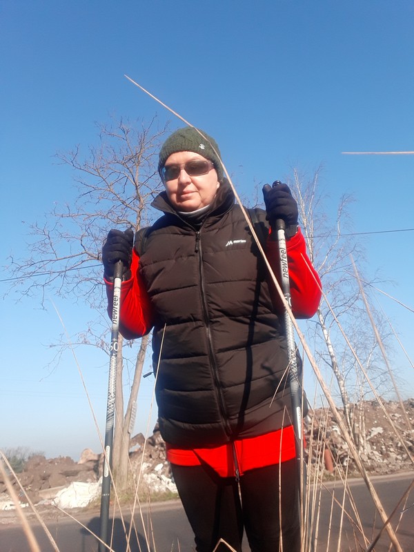 Kobieta w okularach w czerwonej bluzie i czarnej kamizelce, zielonej czapce, stoi oparta o kije nordic walking na tle niebieskiego nieba