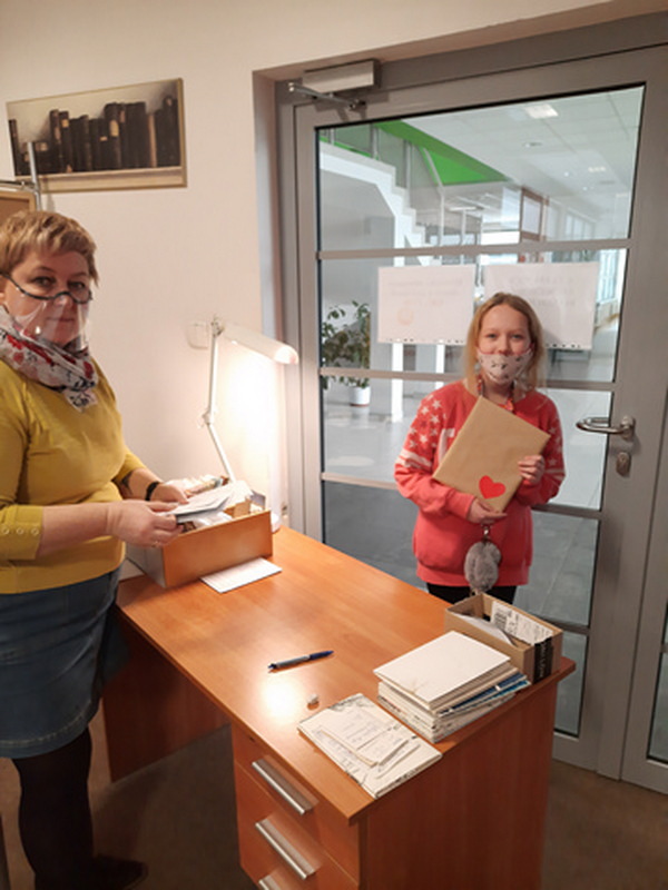 bibliotekarz stoi po jednej stronie biurka, po drugiej stronie stoi dziewczynka w maseczce ochronnej, która trzyma w ręku książkę zawiniętą w szary papier z naklejonym czerwonym serduszkiem