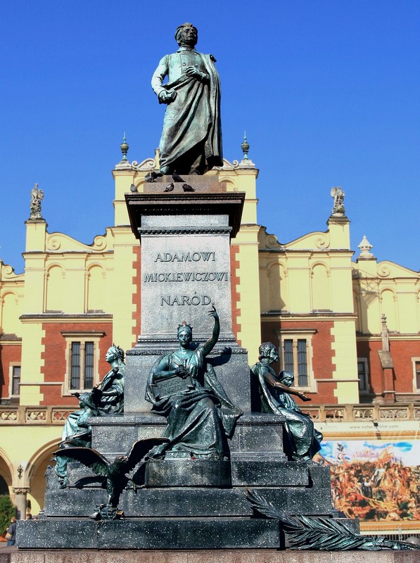 na pierwszym planie pomnik Adama Mickiewicza w Krakowie. Na pomniku napis: Adamowi Mickiewiczowi naród. Za nim widoczne kamienice