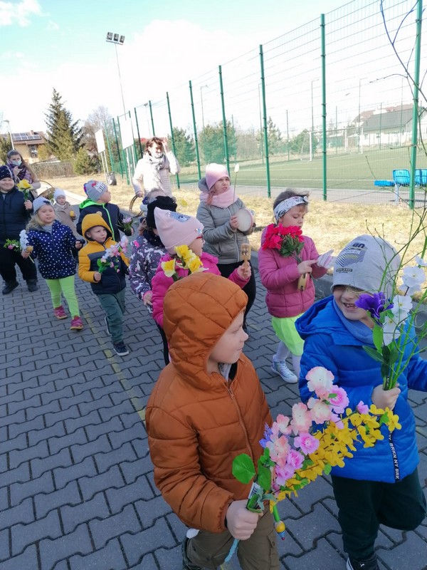 grupa dzieci z klasy 0 ubrana w kurtki i czapki spaceruje wokół szkoły z okazji powitania wiosny. Dzieci mają kwiatki w rękach