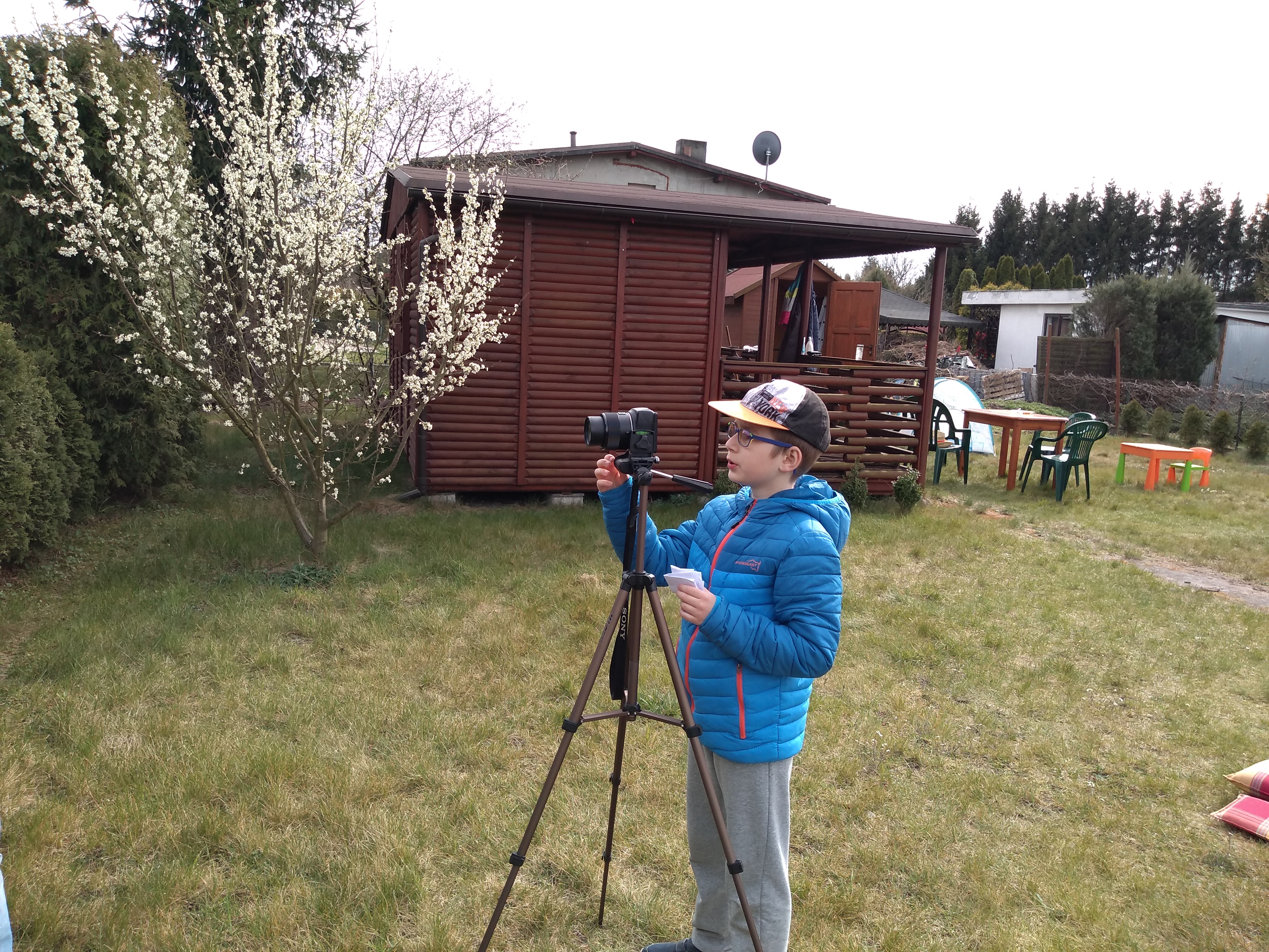 chłopiec stoi w ogrodzie przed aparatem fotograficznym stojącym na statywie. Na drugim planie widać drewnianą altanę i kwitnące drzewo owocowe