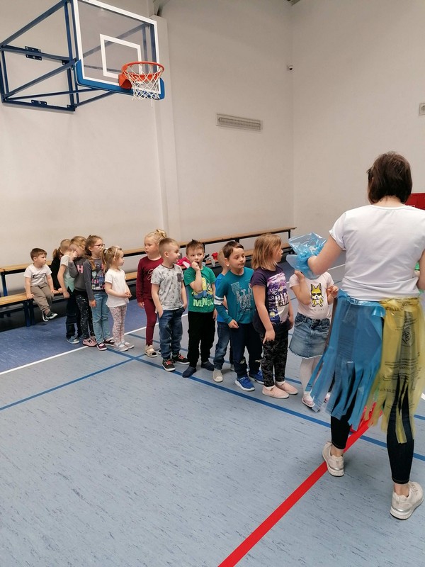 sala gimnastyczna, dzieci stoją w rzędzie, przed nimi wychowawczyni ubrana w turkusowy fartuch