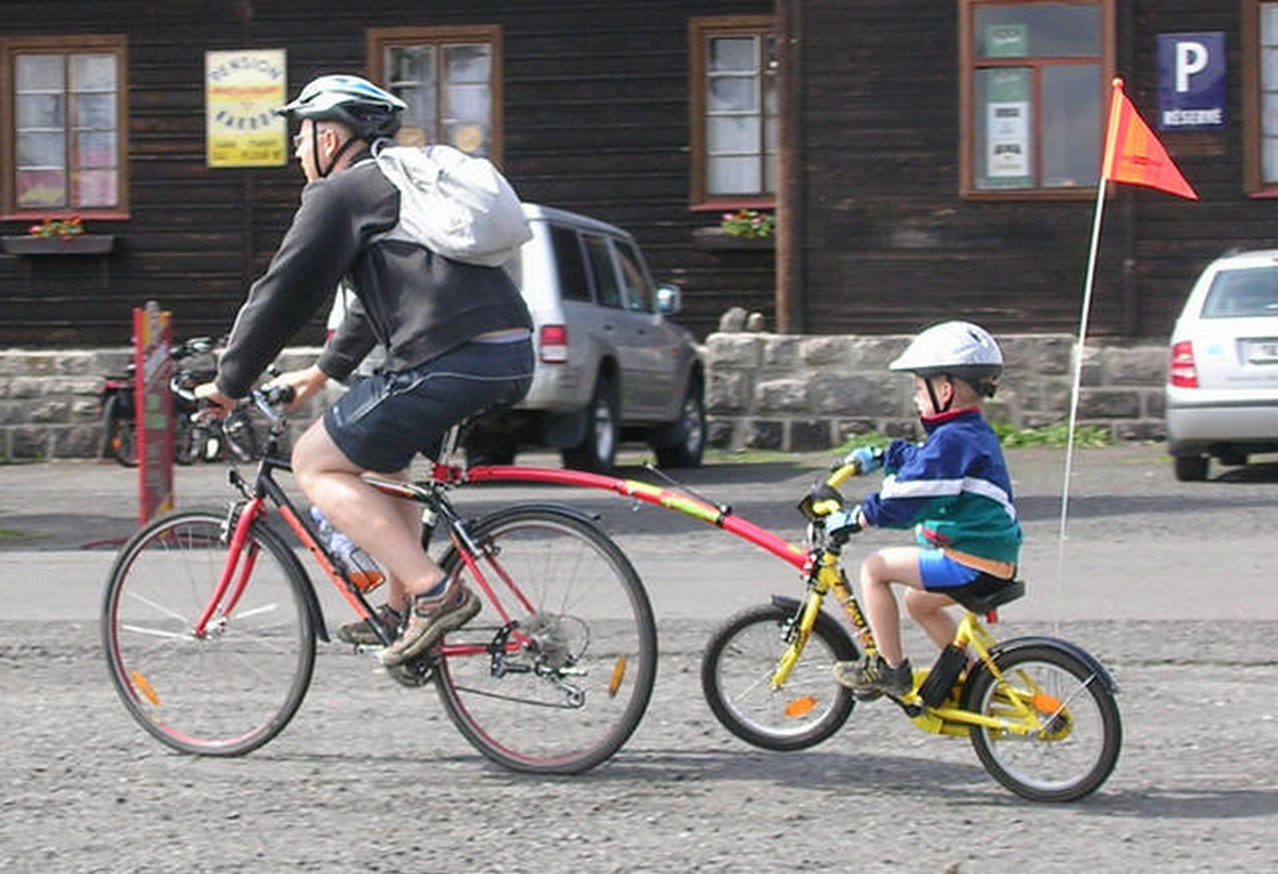 jadący rowerzysta, a za nim doczepiony dziecięcy rowerek z dzieckiem w kasku